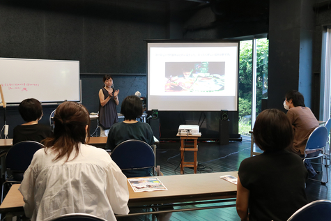 A view from the back of the classroom of a lecture being given while looking at a screen projected by a projector in a dark room. A woman is lecturing next to the screen, and the backs of students sitting at several long desks and metal chairs are visible as they listen to the lecture